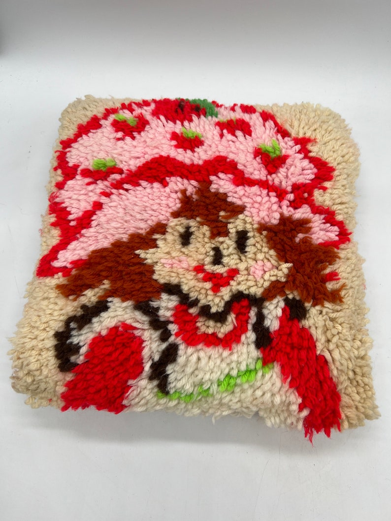 Op de afbeelding: Een vintage naaldviltkussen met een cartoonfiguur met rood haar, een roze hoed en een rood hart. Het figuur is omringd door wit, rood en groen garen.