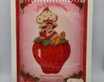 Könnte beinhalten: Ein gerahmtes Bild von Erdbeer-Torte, einer Comicfigur, die auf einer großen roten Erdbeere sitzt. Das Bild hat einen rosa Hintergrund und der Text "Life is the berries!" und "Strawberry Shortcake" sind darauf gedruckt.