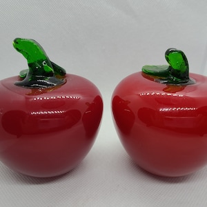 Puede incluir: Dos manzanas de vidrio rojo con tallos de vidrio verde. Las manzanas son brillantes y reflejan la luz.