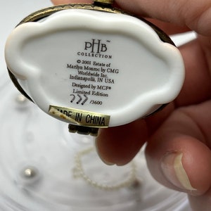 May include: White porcelain trinket box with a gold-tone metal lid and chain. The box has a label with the text "PHB Collection 2001 Estate of Marilyn Monroe by CMG Worldwide Inc. Indianapolis, IN USA Designed by MCF&reg; Limited Edition 777 /3600 MADE IN CHINA".