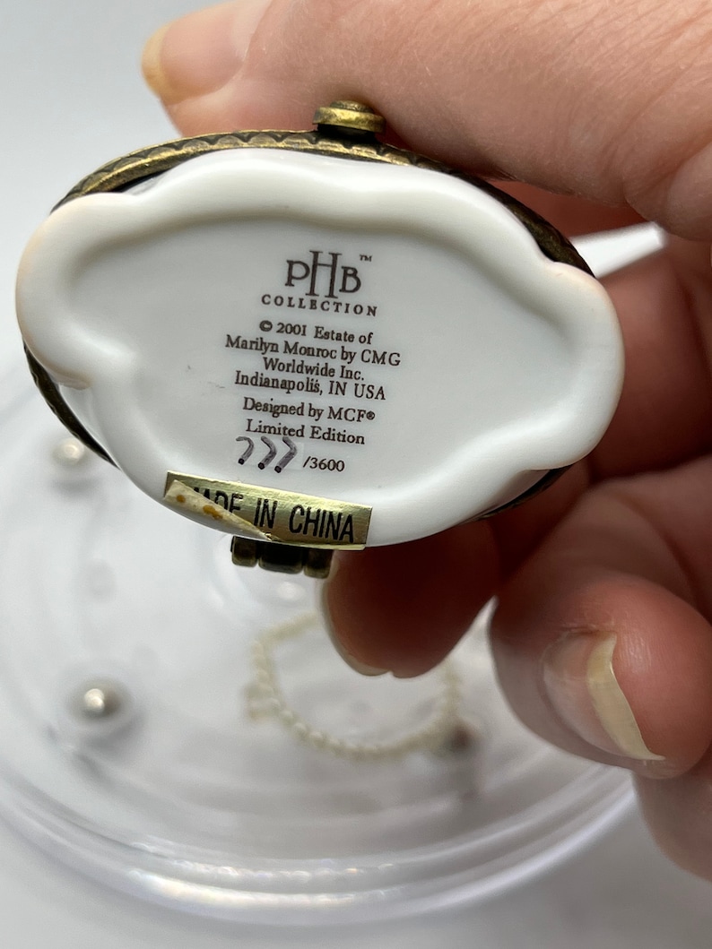 May include: White porcelain trinket box with a gold-tone metal lid and chain. The box has a label with the text "PHB Collection 2001 Estate of Marilyn Monroe by CMG Worldwide Inc. Indianapolis, IN USA Designed by MCF&reg; Limited Edition 777 /3600 MADE IN CHINA".