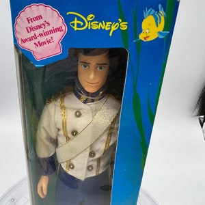 Pode incluir: Uma boneca de pl&aacute;stico do Pr&iacute;ncipe Eric do filme da Disney A Pequena Sereia, com seu uniforme azul e branco. A boneca est&aacute; embalada em uma caixa azul com uma imagem de um peixe amarelo e as palavras "Disney's" e "From Disney's Award-Winning Movie!"