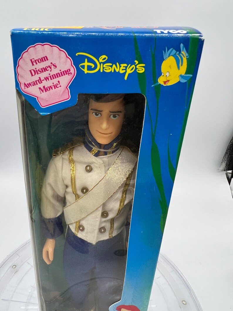 Pode incluir: Uma boneca de pl&aacute;stico do Pr&iacute;ncipe Eric do filme da Disney A Pequena Sereia, com seu uniforme azul e branco. A boneca est&aacute; embalada em uma caixa azul com uma imagem de um peixe amarelo e as palavras "Disney's" e "From Disney's Award-Winning Movie!"