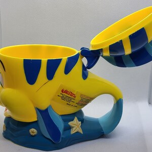 Puede incluir: Taza de pl&aacute;stico amarilla y azul con forma de Flounder de La Sirenita. La taza tiene una tapa y un asa. La taza tiene una pegatina en el lateral que dice "Walt Disney World" y "World of Fun".