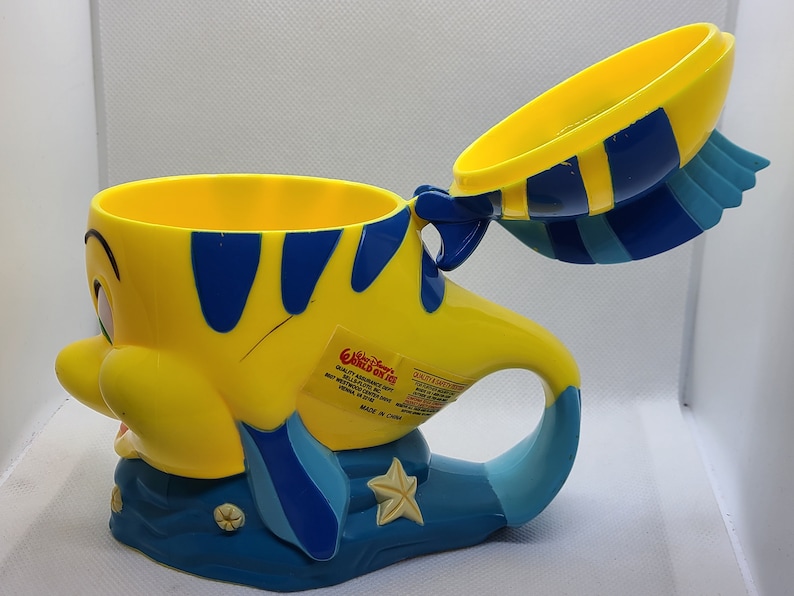 Puede incluir: Taza de pl&aacute;stico amarilla y azul con forma de Flounder de La Sirenita. La taza tiene una tapa y un asa. La taza tiene una pegatina en el lateral que dice "Walt Disney World" y "World of Fun".