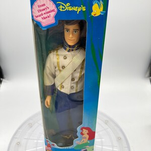 Pode incluir: Uma boneca de pl&aacute;stico do Pr&iacute;ncipe Eric de A Pequena Sereia da Disney, com seu uniforme azul e branco. A boneca est&aacute; embalada em uma caixa azul com uma imagem de Ariel na parte inferior. A caixa diz "From Disney's Award-winning Movie!" e "The Little Mermaid".
