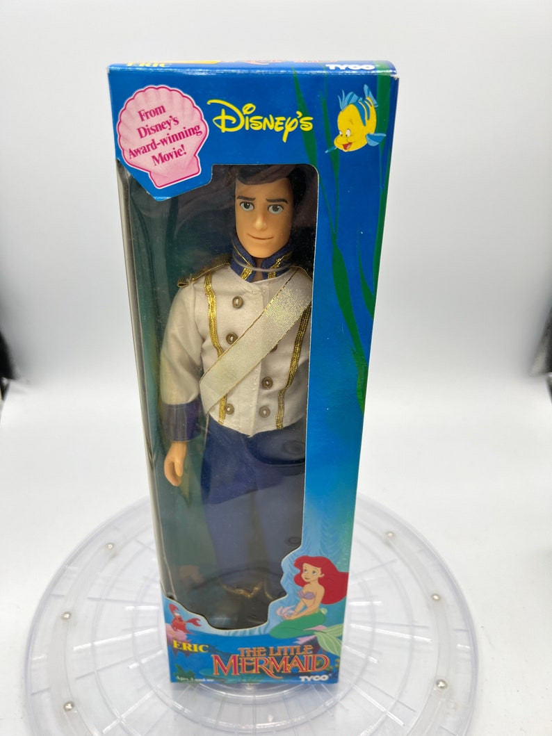 Pode incluir: Uma boneca de pl&aacute;stico do Pr&iacute;ncipe Eric de A Pequena Sereia da Disney, com seu uniforme azul e branco. A boneca est&aacute; embalada em uma caixa azul com uma imagem de Ariel na parte inferior. A caixa diz "From Disney's Award-winning Movie!" e "The Little Mermaid".