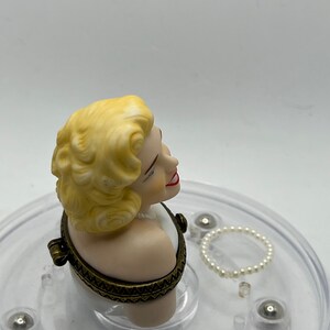 May include: A porcelain bust of a woman with blonde hair, wearing a white dress, with a gold-toned hinged lid. The bust is on a clear plastic base with a pearl necklace.