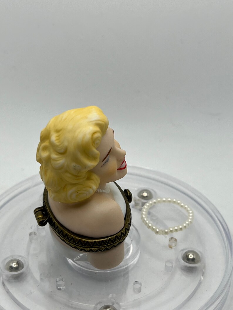 May include: A porcelain bust of a woman with blonde hair, wearing a white dress, with a gold-toned hinged lid. The bust is on a clear plastic base with a pearl necklace.