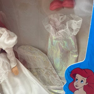 May include: A white wedding dress with a lace trim and a pink bow. The dress is packaged in a blue box with a cartoon image of Ariel from The Little Mermaid.