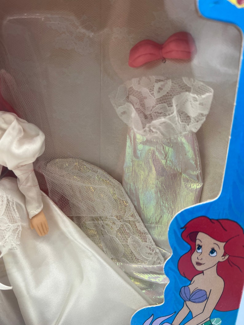May include: A white wedding dress with a lace trim and a pink bow. The dress is packaged in a blue box with a cartoon image of Ariel from The Little Mermaid.