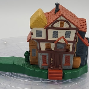 Może przedstawiać: Miniaturowy plastikowy domek z dachem ze słomy, brązowymi ceglanymi ścianami i szarą kamienną fasadą. Domek ma wiele okien i małe drzwi. Domek stoi na zielonej plastikowej podstawie z małą brązową łódką.