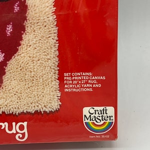 Puede incluir: Una caja roja con una alfombra blanca en su interior. La caja tiene el texto "Craft Master" y "Item No. 35102". El texto en la caja dice "Set contains: Pre-printed canvas for 20" x 27" rug, acrylic yarn and instructions."