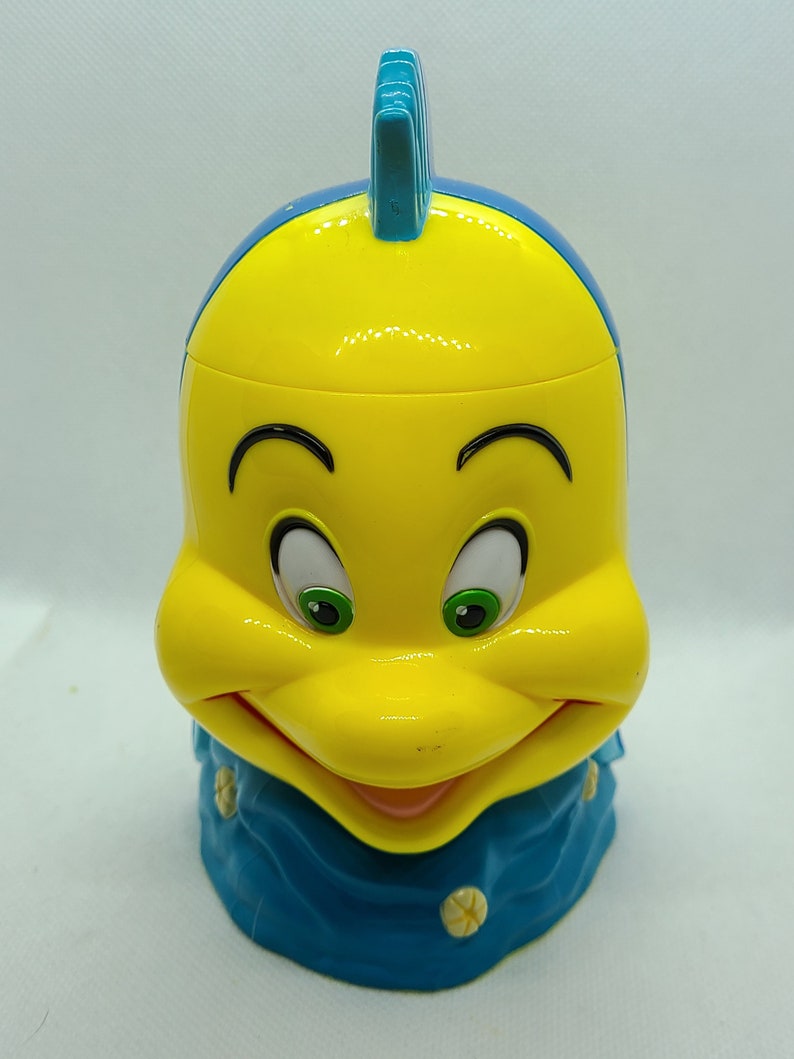 Puede incluir: Figurita de pl&aacute;stico amarilla y azul de un pez de dibujos animados con una cara sonriente. El pez tiene una aleta azul en la parte superior de la cabeza y una cola azul.