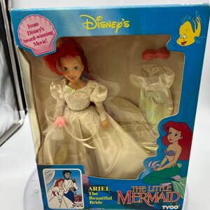 May include: A plastic doll of Ariel from Disney's The Little Mermaid, dressed in a white wedding gown with a pink flower. The doll is packaged in a blue box with a picture of Ariel and the text "Ariel The Beautiful Bride" and "The Little Mermaid".