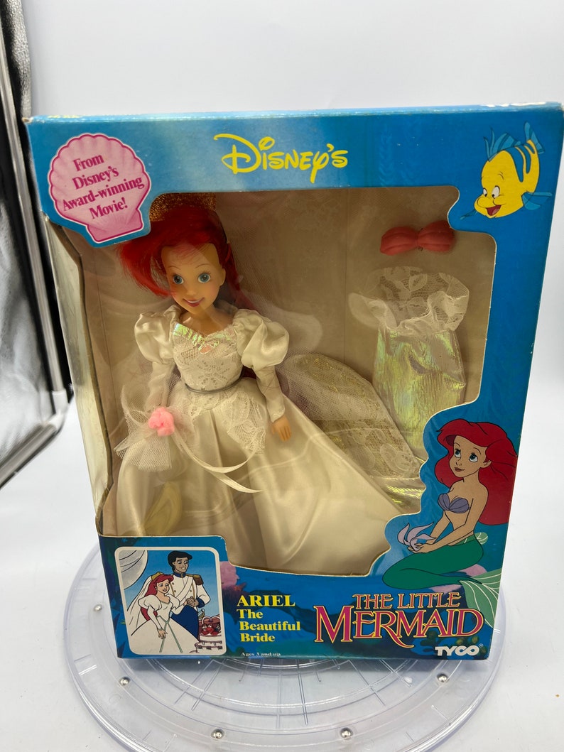 May include: A plastic doll of Ariel from Disney's The Little Mermaid, dressed in a white wedding gown with a pink flower. The doll is packaged in a blue box with a picture of Ariel and the text "Ariel The Beautiful Bride" and "The Little Mermaid".