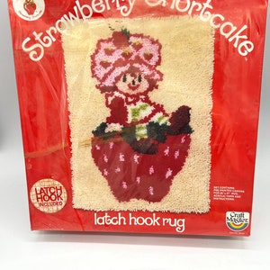 Puede incluir: Un kit de alfombra de gancho de cierre con Strawberry Shortcake. El kit incluye lienzo preimpreso, hilo acr&iacute;lico e instrucciones. "Latch Hook Included" est&aacute; impreso en un c&iacute;rculo rojo.