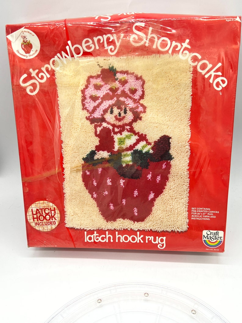 Puede incluir: Un kit de alfombra de gancho de cierre con Strawberry Shortcake. El kit incluye lienzo preimpreso, hilo acr&iacute;lico e instrucciones. "Latch Hook Included" est&aacute; impreso en un c&iacute;rculo rojo.