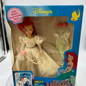 May include: Ariel, the Little Mermaid, is dressed as a bride in a white gown with a pink flower. The box features a cartoon image of Ariel and Prince Eric getting married. The box also includes the text "Ariel The Beautiful Bride" and "The Little Mermaid".