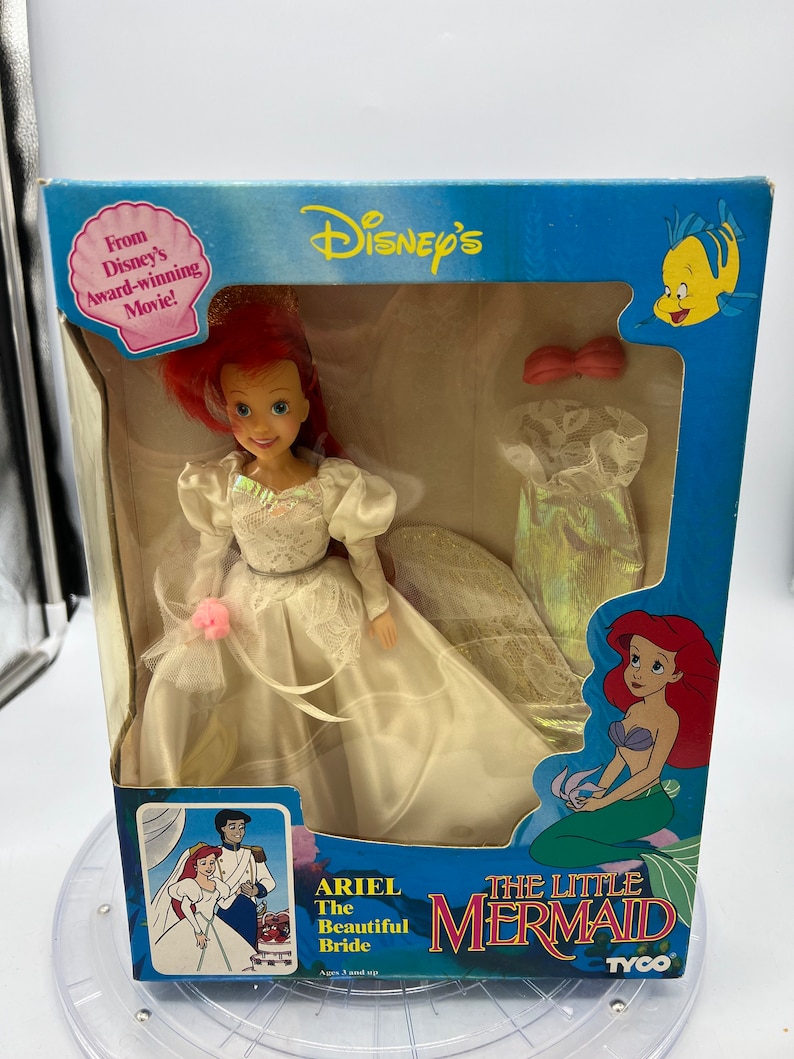 May include: Ariel, the Little Mermaid, is dressed as a bride in a white gown with a pink flower. The box features a cartoon image of Ariel and Prince Eric getting married. The box also includes the text "Ariel The Beautiful Bride" and "The Little Mermaid".