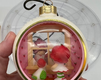 Könnte beinhalten: Ein weißer Weihnachtsbaumschmuck mit goldener Verzierung und einem klaren Fenster. Im Fenster befinden sich zwei Strawberry Shortcake-Figuren und ein Vogel. Der Hintergrund ist rosa mit weißen Erdbeeren.