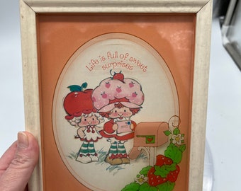 Könnte beinhalten: Ein gerahmtes Bild mit einem pfirsichfarbenen Hintergrund, auf dem zwei Strawberry Shortcake-Figuren neben einem roten Briefkasten stehen. Der Text "Life is full of sweet surprises" ist in einem Kreis um die Figuren geschrieben.
