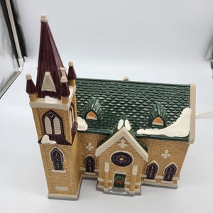 K&ouml;nnte beinhalten: Eine Miniatur-Kirchenfigur mit einem gr&uuml;nen Dach, einem braunen Turm und Buntglasfenstern. Die Kirche ist aus Keramik und hat eine hellbraune Backsteinfassade.