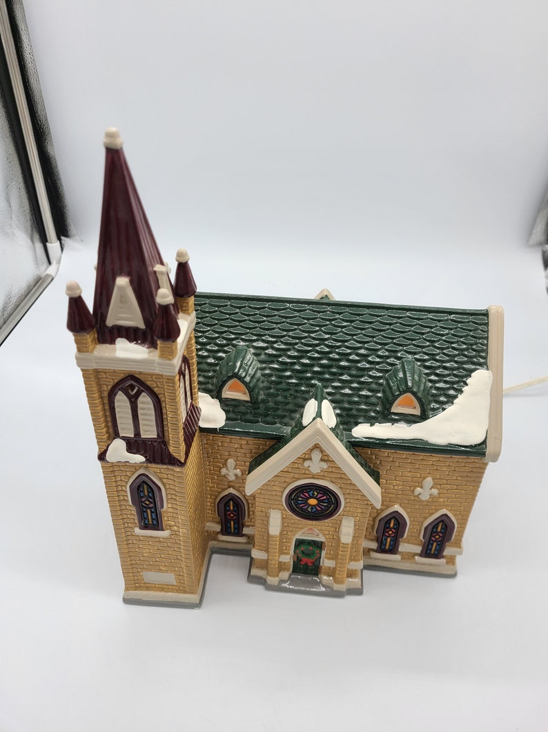 K&ouml;nnte beinhalten: Eine Miniatur-Kirchenfigur mit einem gr&uuml;nen Dach, einem braunen Turm und Buntglasfenstern. Die Kirche ist aus Keramik und hat eine hellbraune Backsteinfassade.
