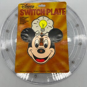 May include: A light brown plastic switch plate featuring a cartoon image of Mickey Mouse with a light bulb above his head. The text "Disney Character Switch Plate" is printed on the packaging. The text "A brilliant idea for adding character to your room!" is also printed on the packaging.