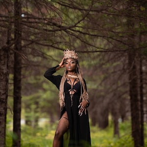 May include: A woman wearing a black maxi dress with a thigh-high slit and a gold crown stands in a forest. She has long blonde braids and is wearing gold jewellery.