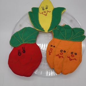 Puede incluir: Tres decoraciones de tela con forma de verduras sonrientes: maíz amarillo con hojas verdes, rábano rojo con hojas verdes y zanahoria naranja con hojas verdes. Cada una tiene rasgos faciales bordados.