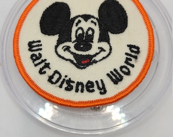 Op de afbeelding: Een witte geborduurde patch met een oranje rand met het gezicht van Mickey Mouse en de tekst "Walt Disney World".