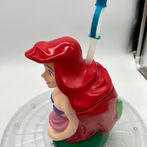 Op de afbeelding: Een rode en groene plastic beker in de vorm van Ariel, de Kleine Zeemeermin, met een blauwe en witte rietje.