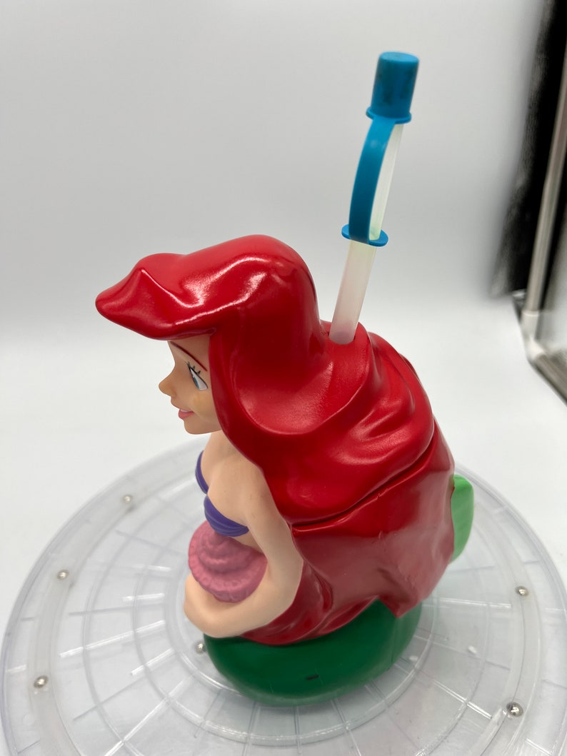 Op de afbeelding: Een rode en groene plastic beker in de vorm van Ariel, de Kleine Zeemeermin, met een blauwe en witte rietje.