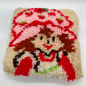 Op de afbeelding: Een vintage naaldviltkussen met een cartoonafbeelding van Strawberry Shortcake. Het kussen is gemaakt van een cr&egrave;mekleurige stof met rood, roze, bruin en groen garen.