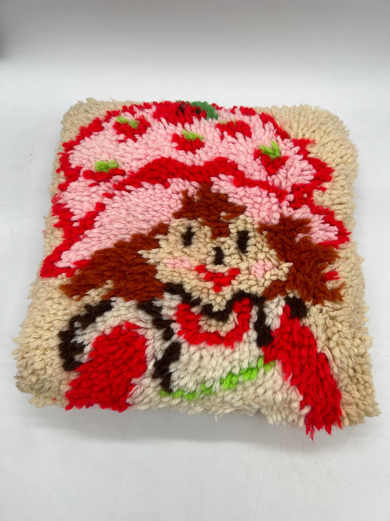 Op de afbeelding: Een vintage naaldviltkussen met een cartoonafbeelding van Strawberry Shortcake. Het kussen is gemaakt van een cr&egrave;mekleurige stof met rood, roze, bruin en groen garen.