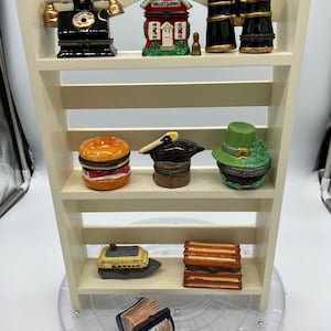 May include: A cream-colored shelf unit displays miniature decorative boxes. The boxes include a black telephone, a red schoolhouse, binoculars, a hamburger, a hat, a green hat, a boat, a stack of books, and an accordion. The shelf unit is on a clear, rotating surface.