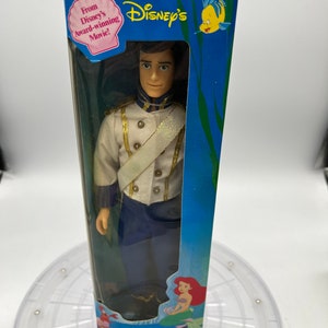 Pode incluir: Uma caixa azul e branca com uma boneca de pl&aacute;stico do Pr&iacute;ncipe Eric de A Pequena Sereia da Disney. A caixa tem uma imagem de Ariel na parte inferior e o texto "A Pequena Sereia" e "Tyco".