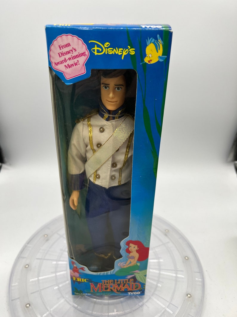 Pode incluir: Uma caixa azul e branca com uma boneca de pl&aacute;stico do Pr&iacute;ncipe Eric de A Pequena Sereia da Disney. A caixa tem uma imagem de Ariel na parte inferior e o texto "A Pequena Sereia" e "Tyco".