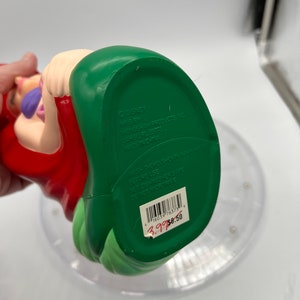 Op de afbeelding: Een groene en rode plastic figuur van Ariel uit De Kleine Zeemeermin. De figuur zit met haar mond open en haar armen uitgestrekt. De onderkant van de figuur heeft de tekst "DISNEY" en "MFG BY VONGGRAM PRODUCTS INC. LABEL PL 22778 MADE IN CHINA". De tekst "WASH THOROUGHLY IN WARM WATER BEFORE USE DISHWASHER SAFE DO NOT MICROWAVE" staat ook op de onderkant van de figuur. Een barcode en een prijskaartje zijn zichtbaar op de onderkant van de figuur.