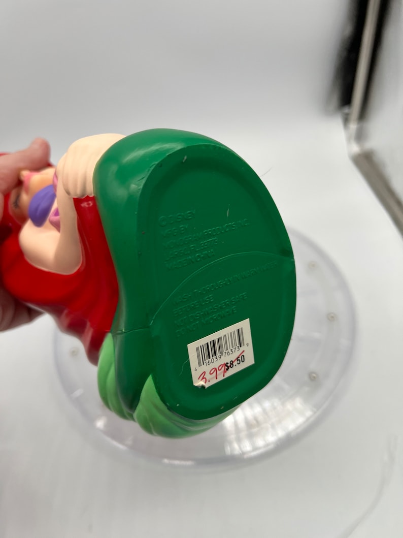 Op de afbeelding: Een groene en rode plastic figuur van Ariel uit De Kleine Zeemeermin. De figuur zit met haar mond open en haar armen uitgestrekt. De onderkant van de figuur heeft de tekst "DISNEY" en "MFG BY VONGGRAM PRODUCTS INC. LABEL PL 22778 MADE IN CHINA". De tekst "WASH THOROUGHLY IN WARM WATER BEFORE USE DISHWASHER SAFE DO NOT MICROWAVE" staat ook op de onderkant van de figuur. Een barcode en een prijskaartje zijn zichtbaar op de onderkant van de figuur.