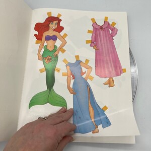 Puede incluir: Un conjunto de mu&ntilde;eca de papel con Ariel de La Sirenita. El conjunto incluye una cola de sirena, un vestido azul con estrellas y una bata de dormir rosa. La mu&ntilde;eca de papel est&aacute; lista para vestirse con los atuendos incluidos.