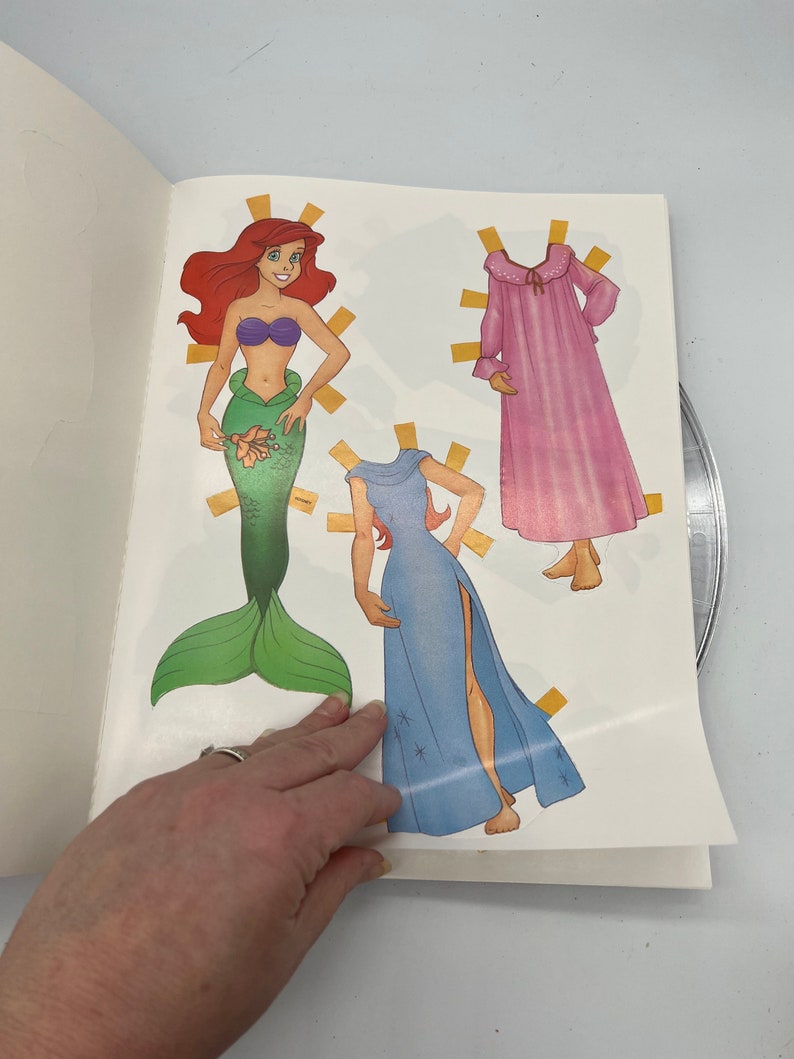 Puede incluir: Un conjunto de mu&ntilde;eca de papel con Ariel de La Sirenita. El conjunto incluye una cola de sirena, un vestido azul con estrellas y una bata de dormir rosa. La mu&ntilde;eca de papel est&aacute; lista para vestirse con los atuendos incluidos.