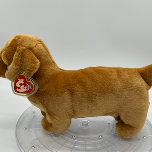 Op de afbeelding: Een bruin pluche speelgoed teckel met een rood en wit Ty-label. Het speelgoed zit op een doorzichtige plastic standaard.