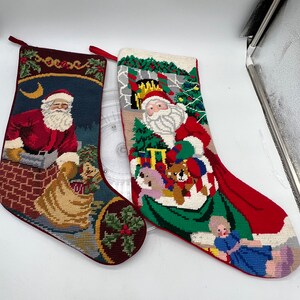 Puede incluir: Dos calcetines navideños con diseños bordados de Papá Noel. Uno muestra a Papá Noel en un tejado, el otro a Papá Noel con regalos y un niño. Ambos calcetines tienen ribetes rojos y lazos para colgar, ideales para la decoración navideña.