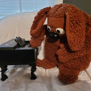 May include: A brown furry toy dog with large eyes and a black nose is sitting on a white surface and playing a small black toy piano.