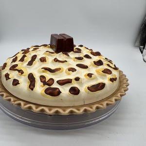 Op de afbeelding: Een keramische taartvorm met een decoratief taartvormig deksel. Het deksel is crèmekleurig met bruine accenten en een donkerbruin chocoladevierkantje bovenop. De taartvorm staat op een heldere, ronde basis.