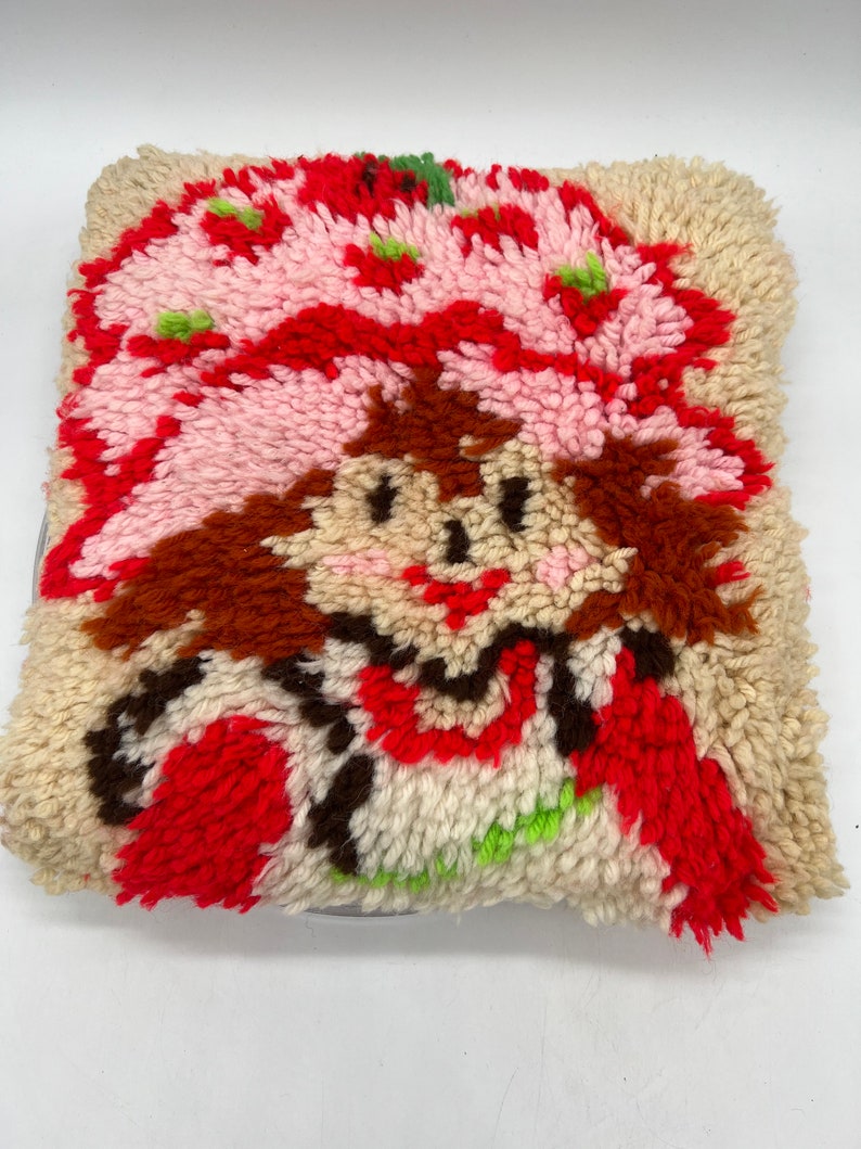 Op de afbeelding: Een vintage-stijl pluche kussen met een cartoon afbeelding van Strawberry Shortcake. Het kussen is gemaakt van een pluche, textuurrijke stof en heeft een rood, roze, wit, bruin en groen kleurenschema.
