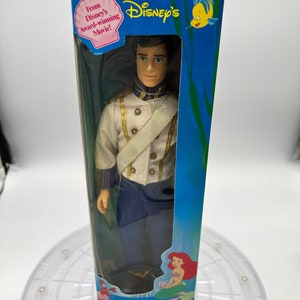 Pode incluir: Uma boneca de pl&aacute;stico do Pr&iacute;ncipe Eric de A Pequena Sereia da Disney. A boneca est&aacute; vestida com um uniforme azul e branco com detalhes dourados. A boneca est&aacute; embalada em uma caixa azul com uma imagem de Ariel na frente. A caixa tamb&eacute;m tem o texto "From Disney's award-winning movie!" e "The Little Mermaid".