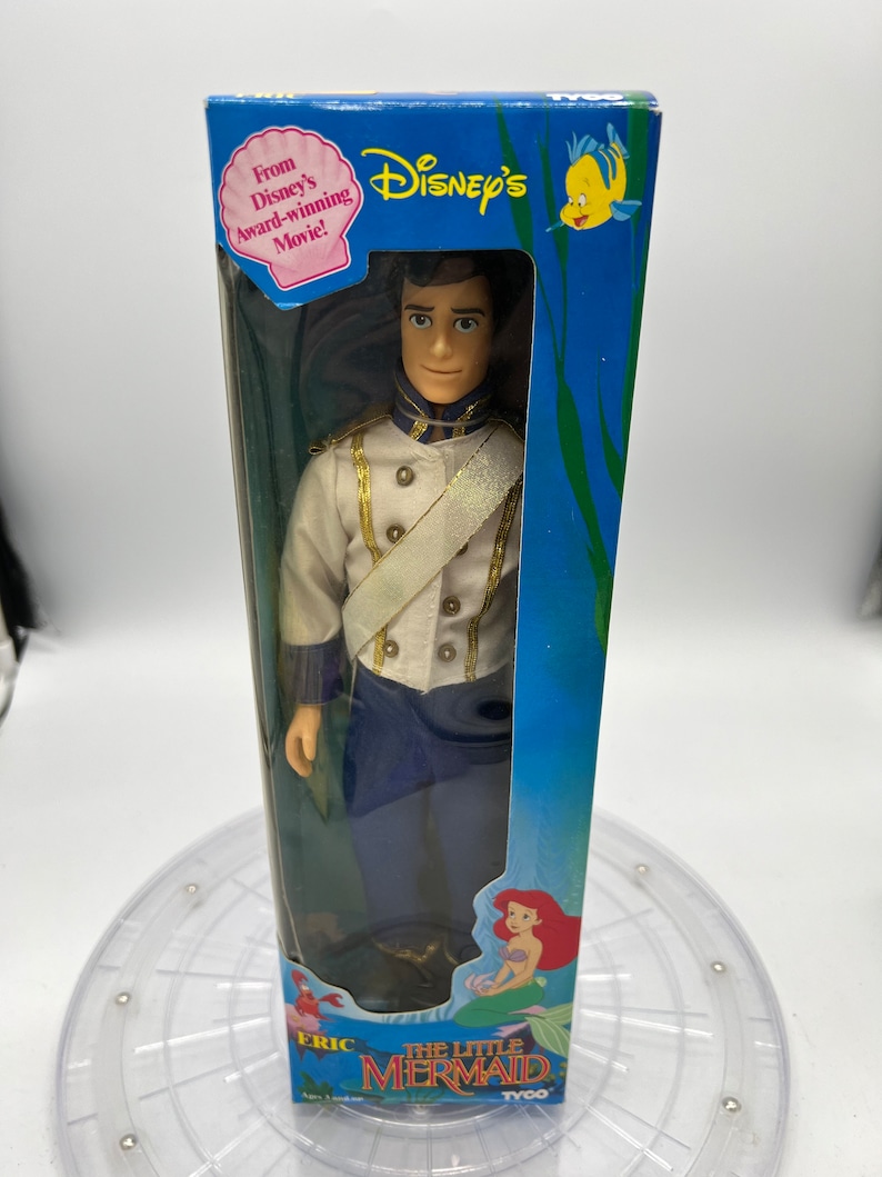 Pode incluir: Uma boneca de pl&aacute;stico do Pr&iacute;ncipe Eric de A Pequena Sereia da Disney. A boneca est&aacute; vestida com um uniforme azul e branco com detalhes dourados. A boneca est&aacute; embalada em uma caixa azul com uma imagem de Ariel na frente. A caixa tamb&eacute;m tem o texto "From Disney's award-winning movie!" e "The Little Mermaid".